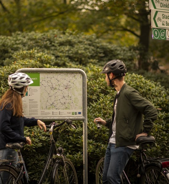Zwei Radfahrer schauen sich eine Karte im Oldenburger Münsterland an. Durchnummerierte Knotenpunkte zeigen, wo es langgeht. 
