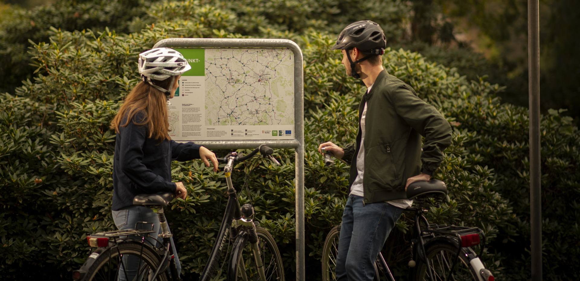 Zwei Radfahrer schauen sich eine Karte im Oldenburger Münsterland an. Durchnummerierte Knotenpunkte zeigen, wo es langgeht. 